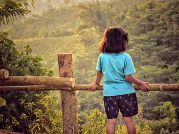 Rear view of boy standing by fence against trees