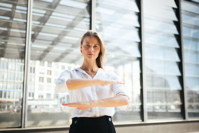Portrait of smiling young woman gesturing equal sign while standing against window