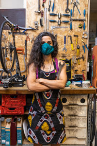 Serious adult ethnic female mechanic in workwear and protective mask holding wrench and looking at camera while standing against weathered workbench with various tools in bicycle service workshop