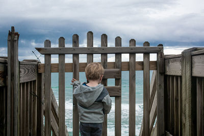 Rear view of boy standing by railing against sky