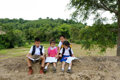 Siblings in school uniforms reading while sitting on field against trees