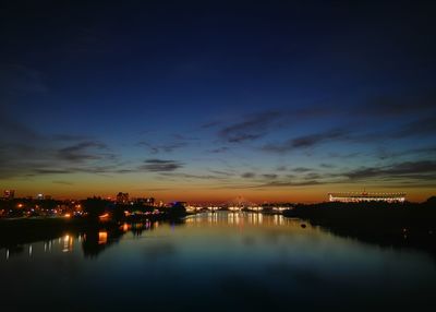 Illuminated city by river against sky at sunset
