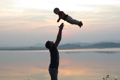 Father throwing son in air by lake against sky during sunset