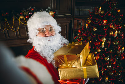 Portrait of man wearing santa claus costume holding christmas presents at home