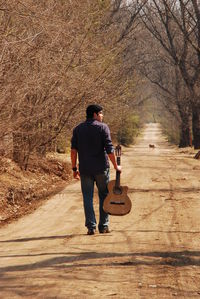 Full length rear view of man walking with guitar on dirt road