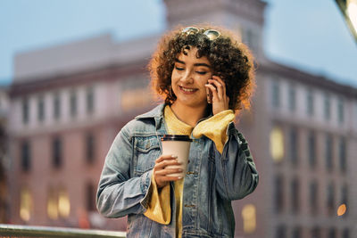Young woman holding coffee cup talking on phone against building