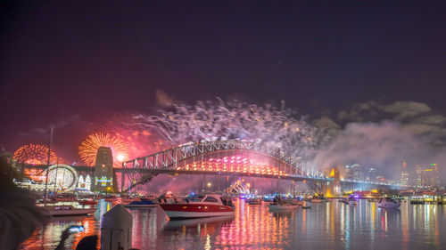 Low angle view of illuminated bridge over river with firework display in background at night