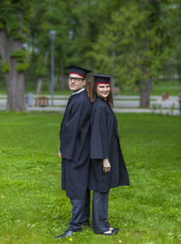 Portrait of friends in graduation gown standing against trees