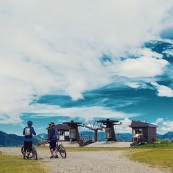 Rear view of bikers with bicycles on dirt road against cloudy sky