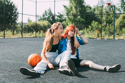 Two girls in sports clothes and with a basketball are chatting, sitting on the playground. sports