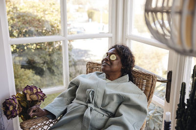 Mature woman with cucumber slices on eyes sitting on chair at home