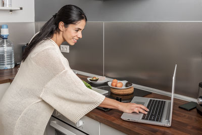 Side view of smiling woman using laptop at kitchen