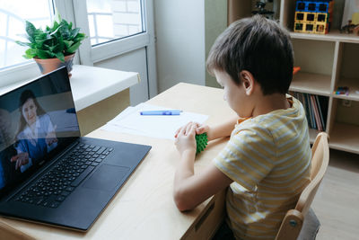 8 years old boy sit by desk with laptop and do exercise with massage ball. smiling woman on screen