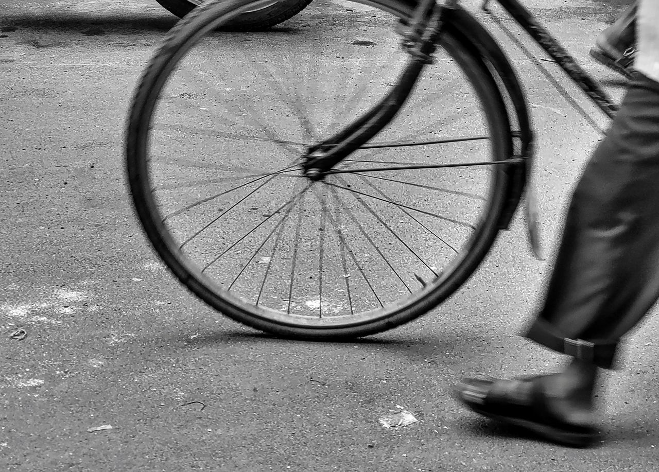 transportation, low section, black, bicycle, human leg, black and white, city, road bicycle, street, vehicle, wheel, monochrome photography, one person, monochrome, mode of transportation, day, white, lifestyles, men, land vehicle, road, motion, limb, outdoors, human limb, footwear, adult, shoe, tarmac, sports equipment, tire, cycling, on the move, bicycle wheel, footpath, standing, blurred motion, activity, pedal, leisure activity