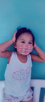 Cute baby girl wearing mask standing on chair against wall