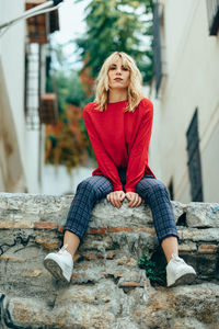 Low angle portrait of young woman sitting on retaining wall in city