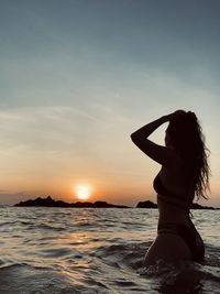 Woman wearing bikini while standing in sea against sky during sunset