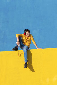 Boy listening to music through headphones while sitting on yellow railing against blue wall
