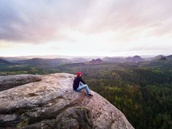 Hiker man in red cap, black sweatshirt and jeans sit. cold morning sun hidden in cloudy sky