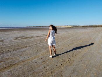 A romantic curly-haired girl walks along the ocean in a light white dress.