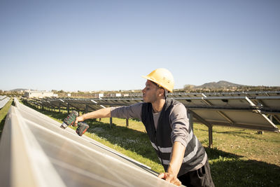 Young male engineer drilling while working near solar panels at power station