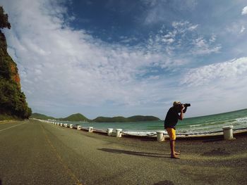 Man photographing through camera while standing at beach against sky