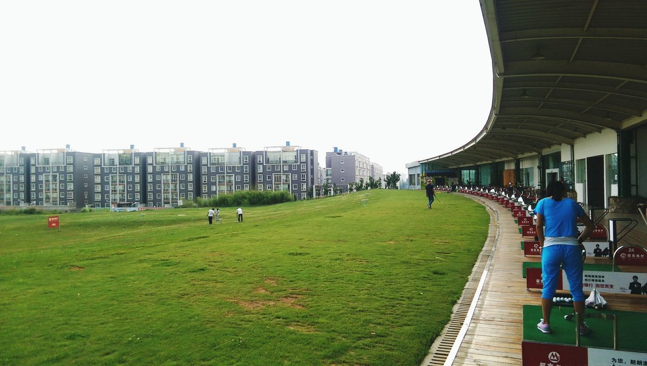 grass, men, person, large group of people, walking, lifestyles, leisure activity, built structure, architecture, building exterior, clear sky, the way forward, city, green color, full length, group of people, mixed age range, park - man made space, day