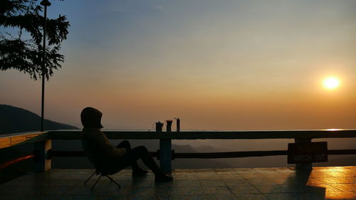 Silhouette man sitting by swimming pool against sky during sunset