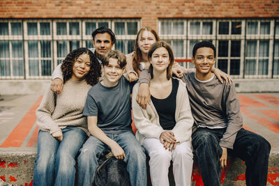 Portrait of smiling male and female students sitting with arms around in high school campus