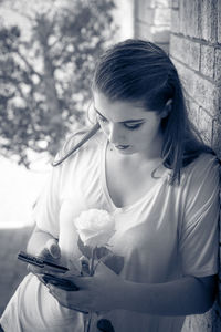 Woman holding rose while using mobile phone against wall