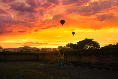 Rear view of woman standing by wall against sky with hot air balloon during sunset