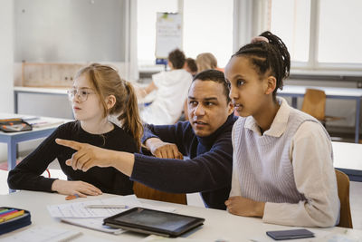 Male teacher pointing by female students sitting at desk in classroom