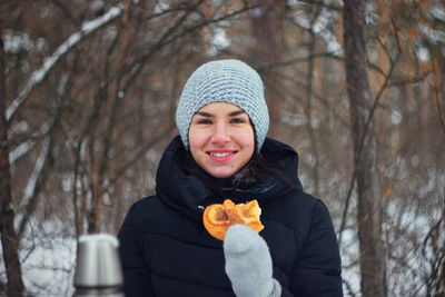 Portrait of smiling young woman holding food while standing against bare trees during winter