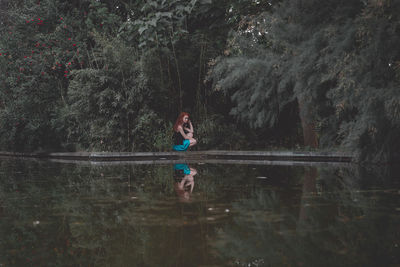 Side view of teenage girl crouching by lake at forest
