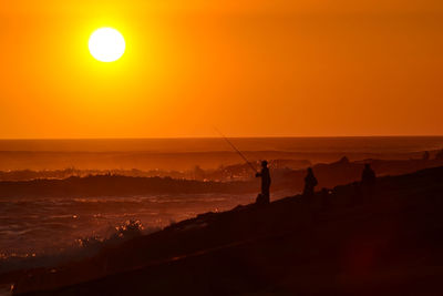 Silhouette man fishing on beach against orange sky