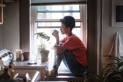 Side view of woman drinking drink while sitting on window sill at home