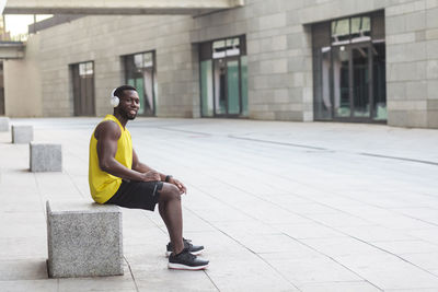 Side view of young man sitting on footpath against building