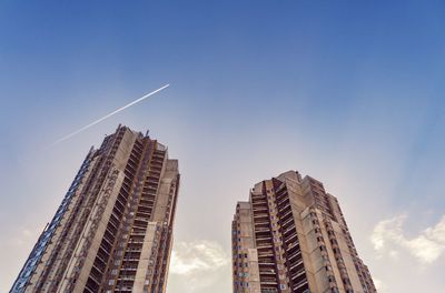 Low angle view of modern buildings against blue sky, plane flying through.