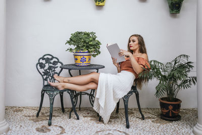 Side view of graceful female in skirt leaning legs on chair while chilling in patio in summer and enjoying interesting story in book at weekend