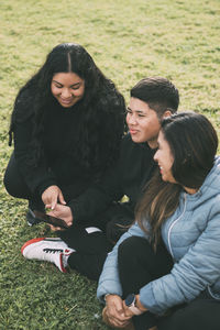 Three people of hispanic-latino ethnicity sitting on the green grass of a park, with smart device