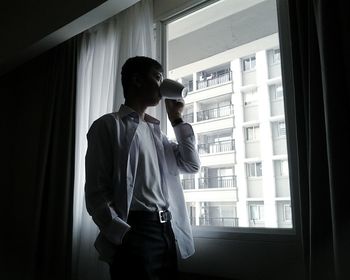 Low angle view of young man drinking coffee by window in apartment