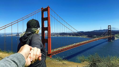 Cropped hand of people with holding hands by bridge against sky