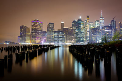 Illuminated modern buildings by river against sky at night