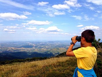 Rear view of boy looking through binoculars at landscape against blue sky