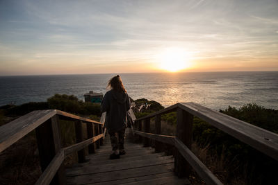 Rear view of man standing on railing against sea during sunset