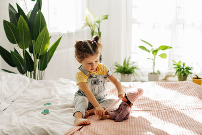 A little girl is sitting on the bed in the bedroom and playing with her doll, tying her scarf