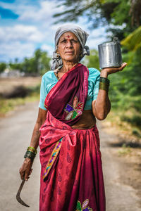 Portrait of mature woman holding container while standing on road against sky