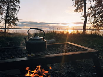 Kettle on bonfire by lake against sky at sunset