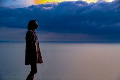 Rear view of woman standing at beach against sky during sunset