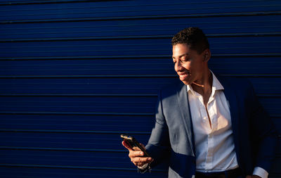 Happy young african american male manager in elegant formal clothes reading message on smartphone and smiling while standing against dark blue wall on street